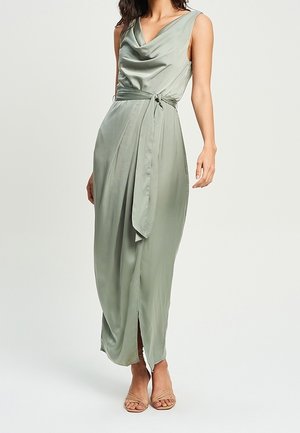 Femme portant une robe portefeuille sans manches vert sauge avec une ceinture à nouer et une fente à l'avant, accompagnée de talons ouverts couleur nude.