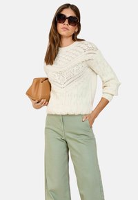 Cremfarbener Strickpullover mit strukturiertem Muster, kombiniert mit hellgrünen weitgeschnittenen Hosen. Das Model hält eine braune Clutch und trägt übergroße Sonnenbrillen.