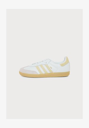 Biele tenisky Adidas Samba s béžovými semišovými pruhmi, záplatou na päte, prekrytím na špičke a gumovou podrážkou na bielom pozadí.