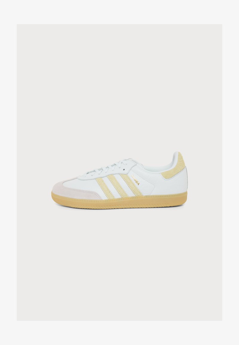 Λευκό αθλητικό παπούτσι Adidas Samba με μπεζ σουέντ ρίγες, πίσω τακούνι, επικάλυψη μύτης και λαστιχένια σόλα σε λευκό φόντο.