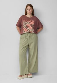 Bruine graphic t-shirt met bloemenprint, gecombineerd met losse, lichtgroene broek. Sandalen met dikke beige platformzolen.