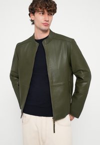 Olijfgroene leren jas met opstaande kraag, rits aan de voorkant en zijzakken. Gladde textuur en getailleerde pasvorm. Donkere trui eronder.