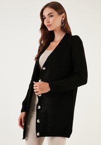 Fekete kardigán texturált kötött anyagból, V-nyakkivágással, hosszú ujjal és nagy ezüstszínű gombokkal az elején.