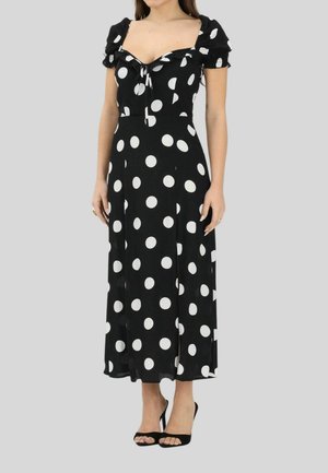 Donna con un vestito midi nero a pois bianchi, maniche a sbuffo, scollo quadrato e tacchi neri aperti, in piedi davanti a uno sfondo semplice.