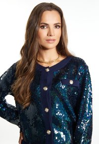 Námornícky kardigán s flitrami, ktorý má okrúhle zlaté gombíky a predné vrecko na hrudi. Hladká textúra a lesklé multikolórové flitre.