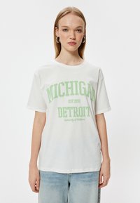 Hvid bomuld t-shirt med grøn tekst, der står "MICHIGAN DETROIT," med en afslappet pasform og korte ærmer. Sammen med lyseblå denim jeans.