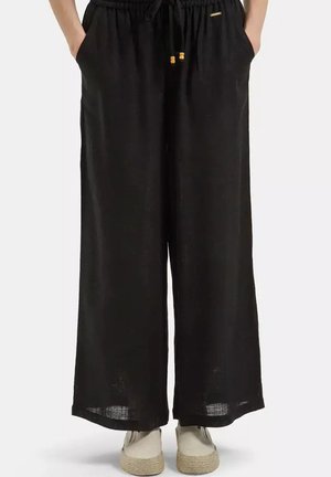 Czarne spodnie o szerokich nogawkach z elastycznym pasem i sznurkiem, ręce w kieszeniach, noszone z beżowymi butami na platformie.
