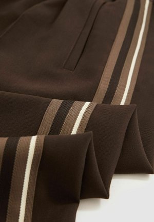 Brauner Stoff mit drei vertikalen Streifen in Beigetönen und Dunkelbraun. Besitzt eine glatte Textur und einen gefalteten Saum mit einer Taschenapplikation.