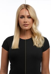 Cheveux blonds séparés en deux : le côté gauche lisse, le côté droit ondulé. Porte un shirt noir à manches courtes. Fond neutre avec une ligne pointillée.