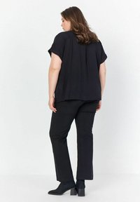 Top nero a maniche corte con vestibilità ampia e pieghe, abbinato a pantaloni neri dritti. L'outfit è completato da stivaletti neri alla caviglia.
