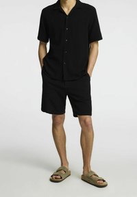 Czarna koszula z krótkim rękawem i kołnierzykiem, zestawiona z czarnymi szortami. Model nosi oliwkowozielone sandały wsuwane z teksturowanym paskiem.