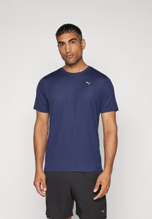 Uomo in maglietta atletica blu Puma e pantaloni corti neri Puma su sfondo chiaro uniforme, in posizione frontale con espressione neutra.