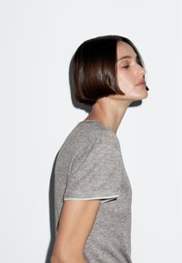 Maglietta grigia a maniche corte con scollatura rotonda e delicati bordi bianchi, indossata da un modello con un taglio a caschetto liscio su uno sfondo bianco.