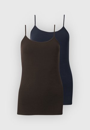 Vrouw met een donkerbruin mouwloos topje, lichtblauwe wijde jeans met grote omslagen, zwarte riem en zwarte puntige schoenen, staand tegen een witte achtergrond.