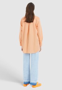 Chemise en coton oversize couleur pêche avec un détail de pli à l'arrière, associée à un jean large bleu clair et des sandales orange vif.