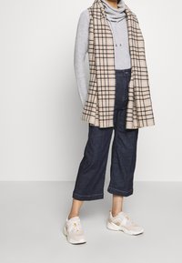 Gray turtleneck, plaid long vest in beige and black, dark blue denim wide-leg pants, and light-colored athletic shoes with accents.