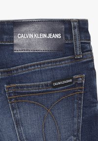 Vaqueros de mezclilla azul oscuro con una etiqueta de cuero con "CALVIN KLEIN JEANS" grabado en blanco. Incluye costuras visibles y detalles de bolsillo.