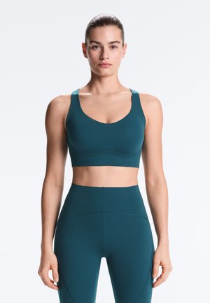 Frau mit zurückgebundenem Haar, die ein türkisfarbenes Sport-BH und hoch taillierte Leggings trägt, steht mit neutralem Gesichtsausdruck frontal zur Kamera.