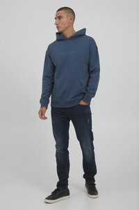 Blaue Kapuzenjacke mit einer frontalen Tasche und verstellbarem Kapuzenzug, kombiniert mit dunklen Denim-Jeans und schwarzen Sneakern. Schlichte Gestaltung mit Logotext.