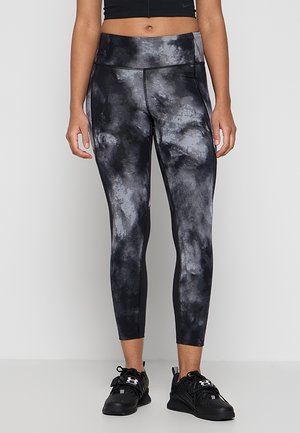 Femme portant des leggings tie-dye noirs et gris, un top court noir et des chaussures de sport noires, debout sur un fond clair uni.