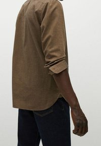 Bruine overhemd met lange mouwen van een textuurstof, gebogen onderkant en opgerolde manchetten, gecombineerd met donkerblauwe jeans. Zichtbare naaddetails en stiksels.