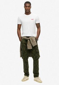 T-shirt bianco in cotone con piccolo logo rosso, pantaloni cargo mimetici verde oliva con tasche, maglione beige legato in vita, sneakers crema.