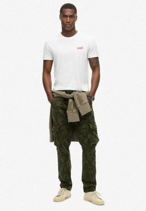 Hvid bomulds t-shirt med lille rødt logo, olivengrøn camouflage cargo-bukser med lommer, beige sweater bundet om livet, cremefarvede sneakers.