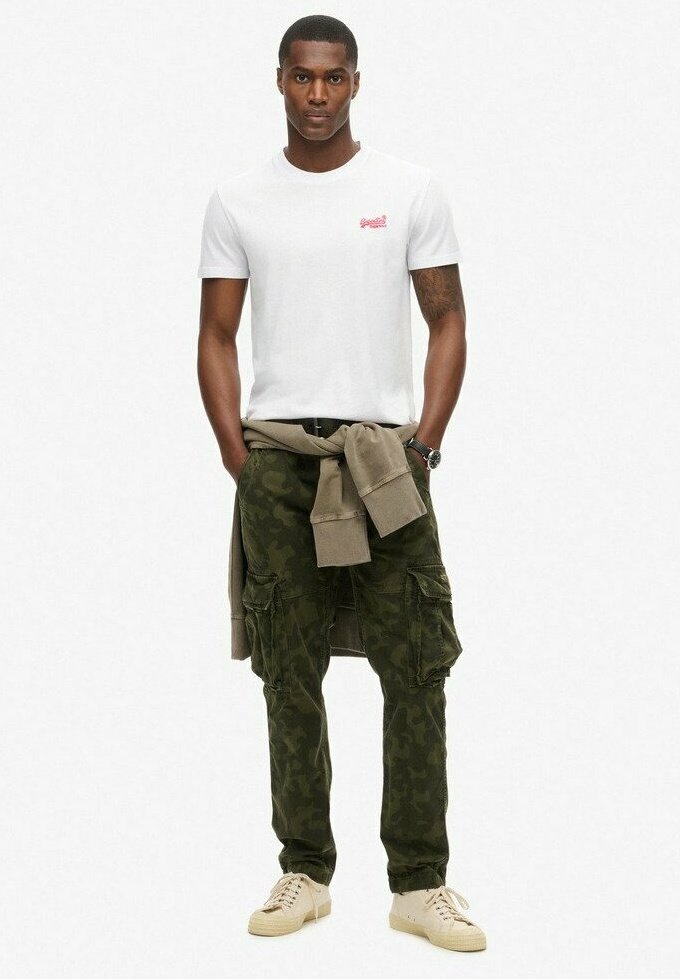 T-shirt bianco in cotone con piccolo logo rosso, pantaloni cargo mimetici verde oliva con tasche, maglione beige legato in vita, sneakers crema.