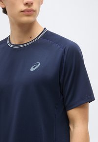 Άτομο που φοράει μπλε σκούρο αθλητικό μπλουζάκι με κοντά μανίκια, ριγωτό γιακά και μικρό ασημένιο λογότυπο ASICS στο στήθος.