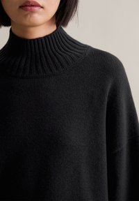 Μαύρο πλεκτό πουλόβερ με ψηλό, ριχτό γιακά; χαλαρή εφαρμογή; λεία υφή; διαθέτει μανίκια ράγκλαν και απλό, μινιμαλιστικό σχέδιο.