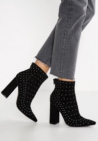 Schwarze Wildleder-Stiefeletten mit spitz zulaufender Zehenpartie und Blockabsatz, verziert mit weißen Nietenakzenten, kombiniert mit ausgefransten grauen Jeans.