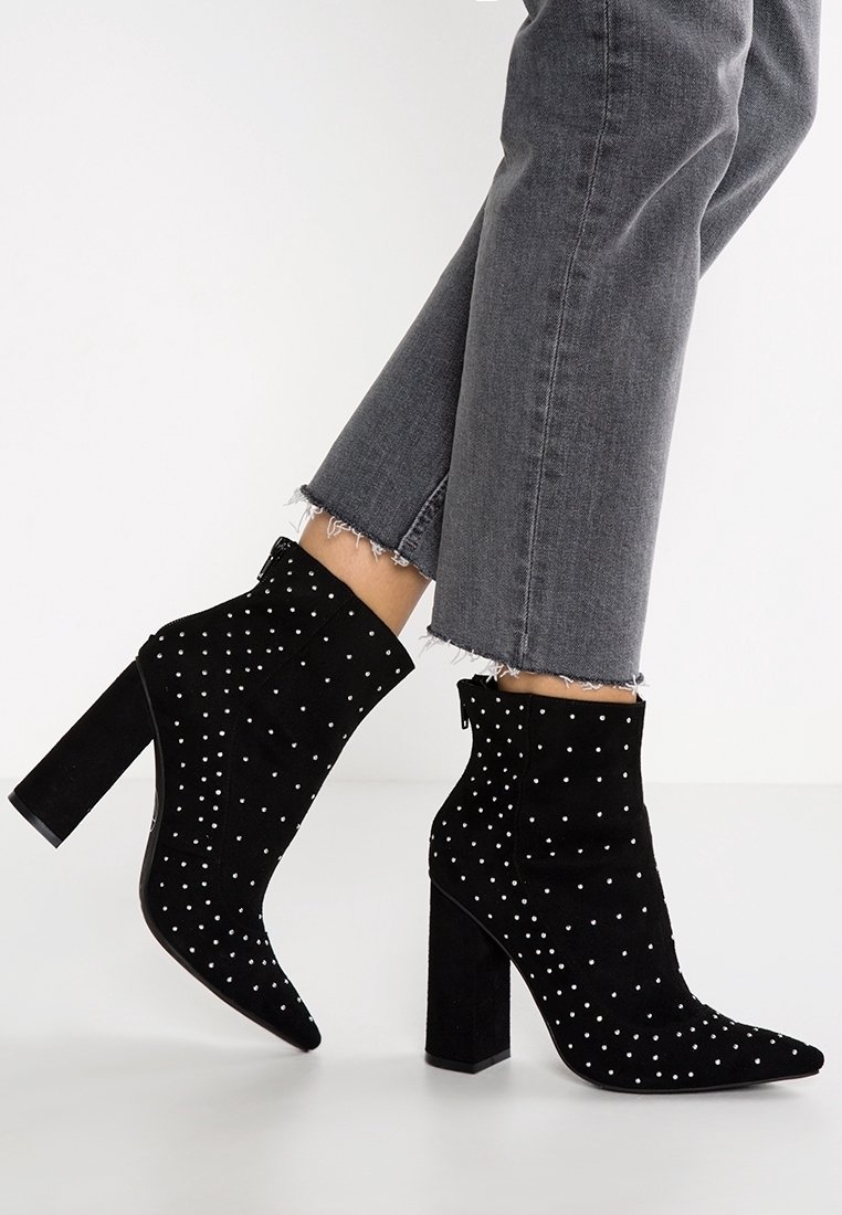 Schwarze Wildleder-Stiefeletten mit spitz zulaufender Zehenpartie und Blockabsatz, verziert mit weißen Nietenakzenten, kombiniert mit ausgefransten grauen Jeans.