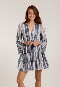 Weißes Kleid mit marineblauen geometrischen Mustern, V-Ausschnitt, Quastenbindung und weit ausgestellten Ärmeln; verfügt über gestufte Rüschendetails am Rock.