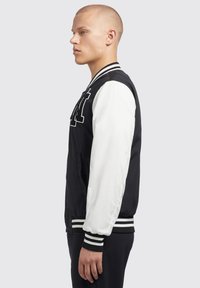 Zwart-wit varsityjack met geribbelde kraag, textuurstof en gestreepte manchetten. Heeft een geborduurd letter op de borst.