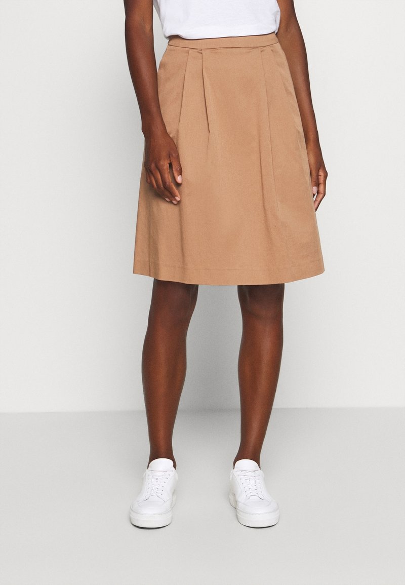Beige A-line nederdel med plisseringer ved livet, lavet af glat stof. Modellen har hvide sneakers på og står foran en neutral baggrund.