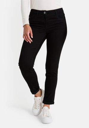 Femme portant un jean slim noir retroussé aux chevilles et des baskets blanches, se tenant avec une jambe légèrement fléchie.