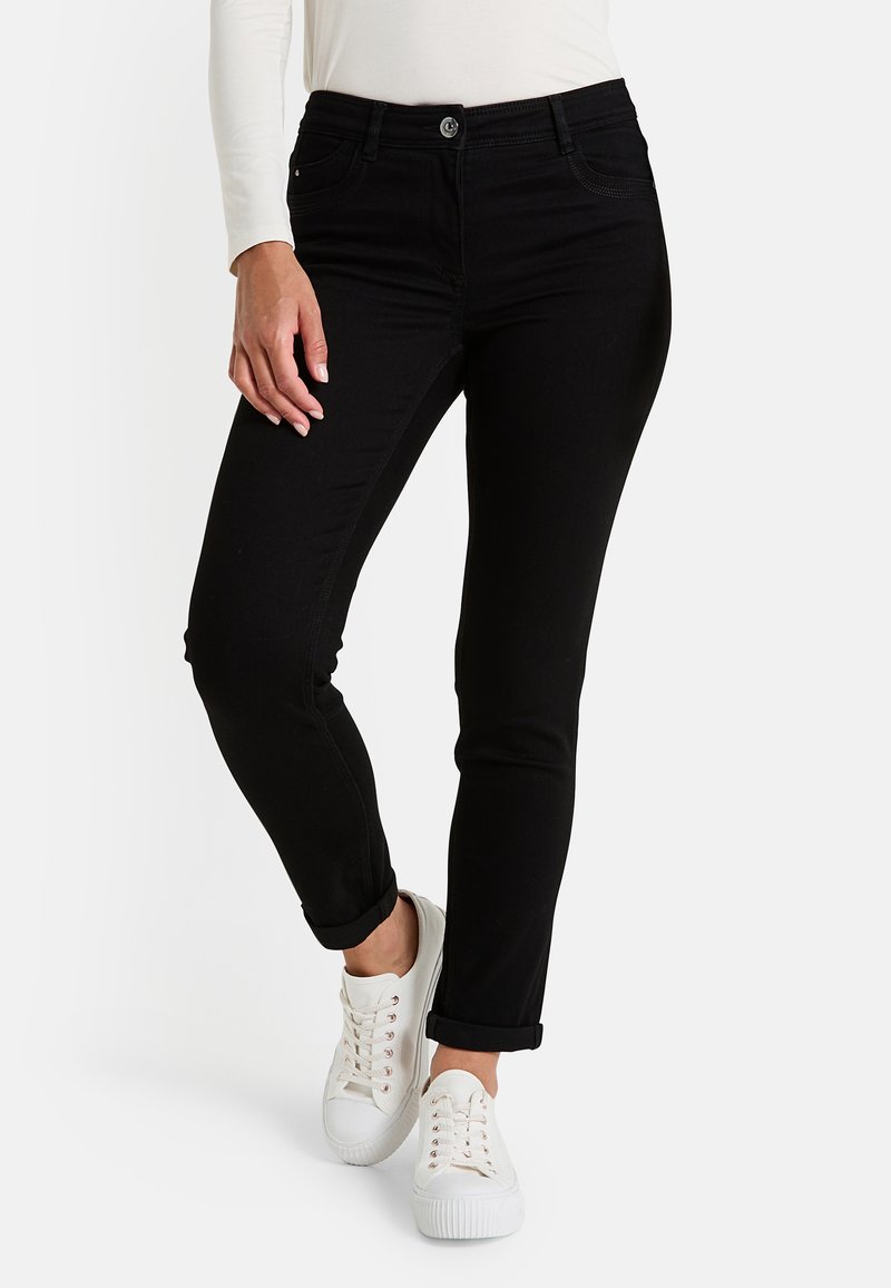 Femme portant un jean slim noir retroussé aux chevilles et des baskets blanches, se tenant avec une jambe légèrement fléchie.