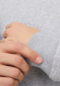 Poignet d'un sweatshirt gris avec une texture côtelée ; main tenant doucement le poignet, mettant en valeur le tissu doux et les détails de couture soignés.