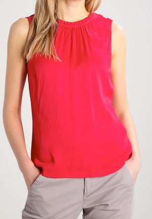 Blouse rouge vif sans manches avec col froncé portée avec un pantalon gris clair, mains posées négligemment dans les poches.