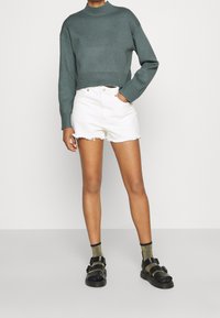 Przycięty zielony sweter z prążkowanym dołem; białe szorty dżinsowe z postrzępionymi brzegami; czarne sandały z dwoma paskami; metaliczne skarpetki.