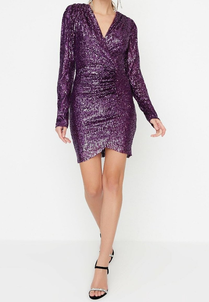 Femme portant une robe portefeuille à manches longues, violette à sequins, avec un ourlet asymétrique et des sandales noires à talons hauts, marchant vers l’avant.