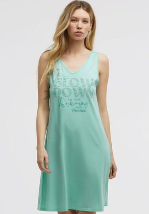 Blonde Frau trägt ein ärmelloses mintgrünes Kleid mit dem Text "Slow Down and start Dreaming again Stories" auf der Vorderseite.