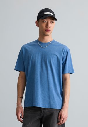 Jonge man die een blauw T-shirt, zwarte pet, zilveren ketting en zwarte spijkerbroek draagt, staand tegen een effen lichtgrijze achtergrond.