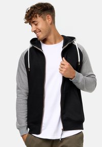Sudadera con cremallera en negro y gris, con tejido texturizado, mangas en contraste, cordones blancos y bolsillos frontales. Se ajusta cómodamente en los hombros.