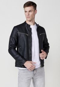 Chaqueta de moto de cuero negro con un corte slim, hombros acolchados, cremalleras plateadas y cuello alto, combinada con una camiseta blanca.