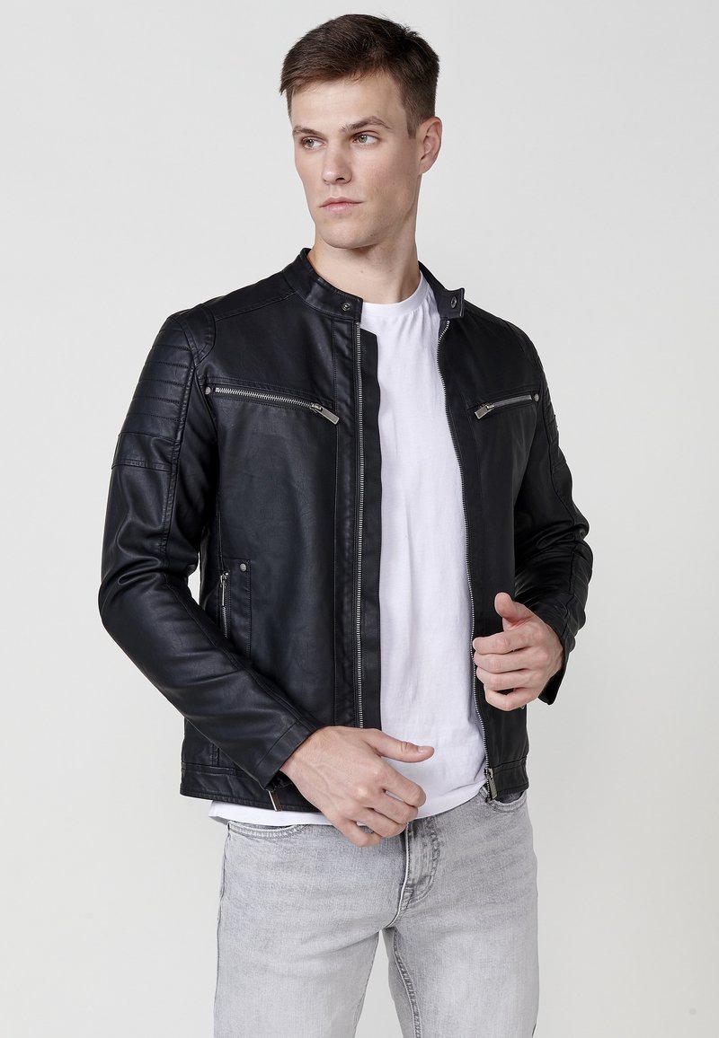Chaqueta de moto de cuero negro con un corte slim, hombros acolchados, cremalleras plateadas y cuello alto, combinada con una camiseta blanca.