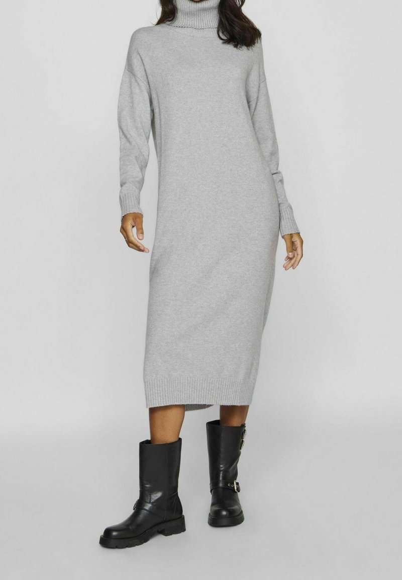 Robe en maille grise à col roulé avec manches longues, longueur mi-mollet, et détails côtelés. Associée à des bottes en cuir noir mi-mollet avec garnitures métalliques.