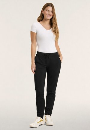 Donna con capelli lunghi e ondulati che sorride, indossa una maglietta bianca a maniche corte, pantaloni neri con coulisse e sneakers bianche con dettagli gialli.