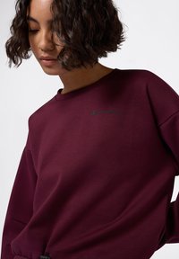 Sudadera burdeos hecha de una tela suave, con un cuello redondo y el logo bordado de "Champion" en verde oscuro en el pecho.