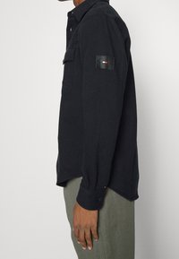 Uomo che indossa una camicia a maniche lunghe di colore blu scuro con tasca sul petto e un logo a patch sul braccio, abbinata a pantaloni color oliva.